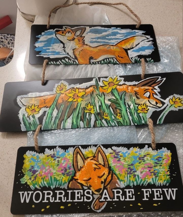 A picture of the sign on my counter, it consists of three 
panels with a cartoony fox enjoying nature. The top panel has the fox looking up to a partly 
cloudy sky, the second has them walking through a field of tall grass and yellow flowers, and
the third has them sleeping in a bed of colorful flowers with the text 'WORRIES ARE FEW'